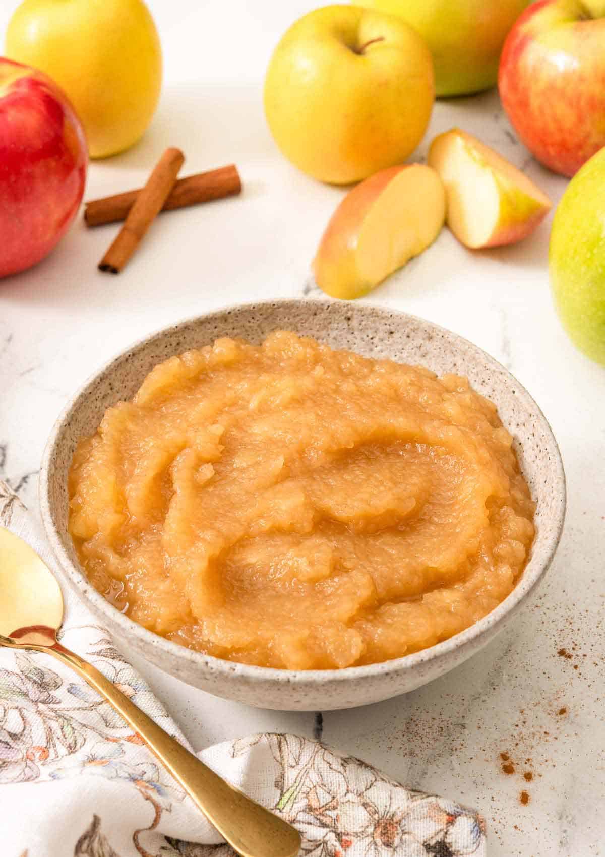 A bowl of apple sauce with fresh apples, some cut, in the background.