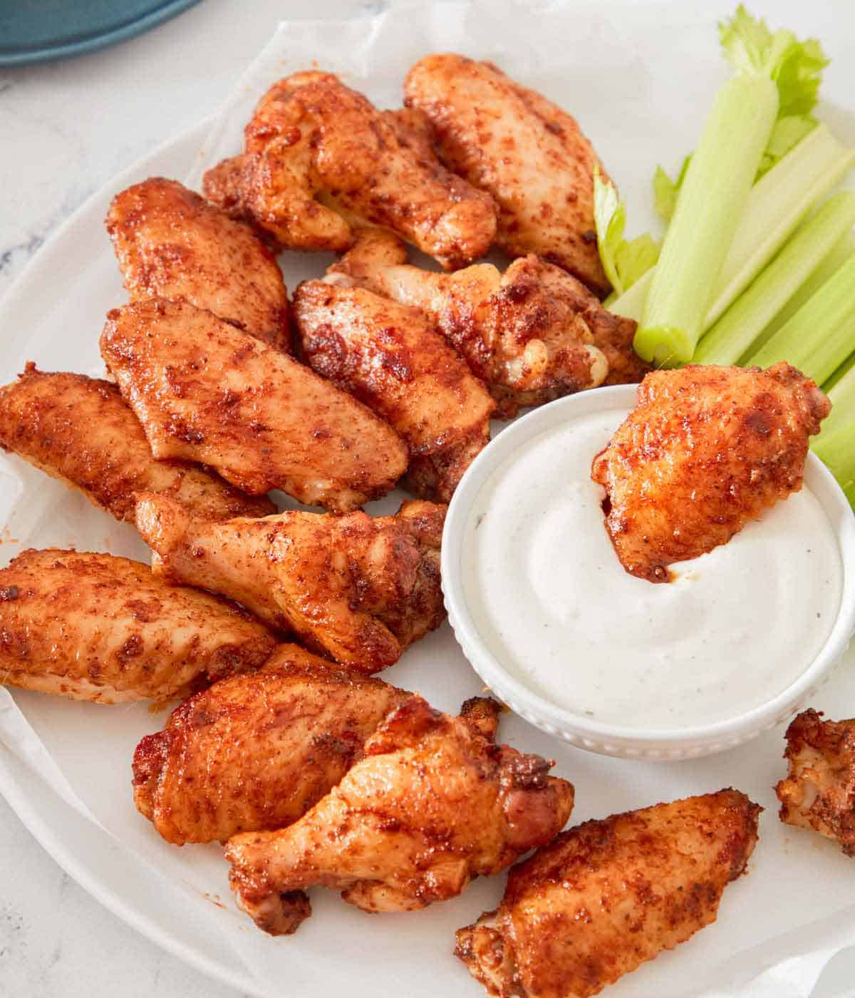 A serving plate with multiple chicken wings and celery with one wing in a bowl of dip.