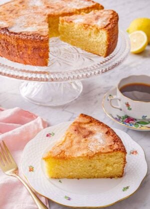 A slice of olive oil cake on a plate in front of the cut cake on a cake stand.