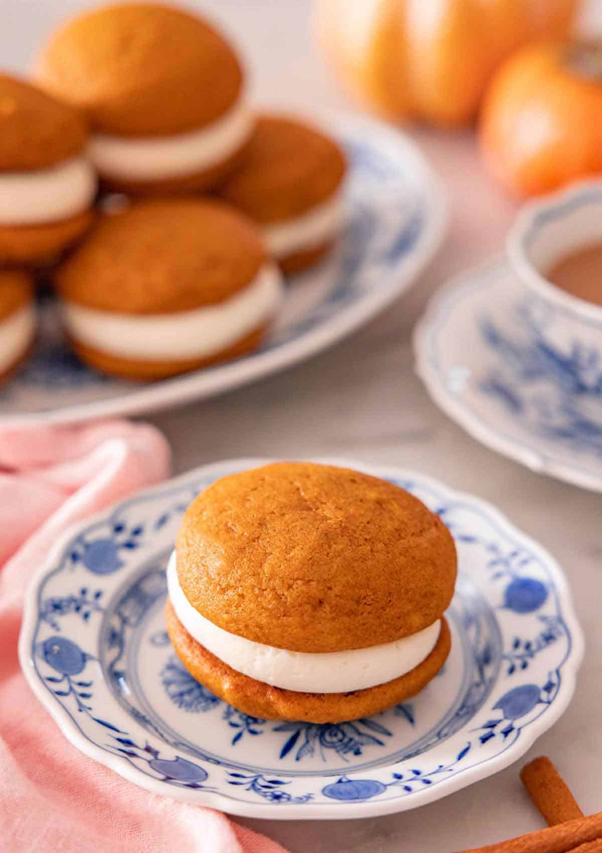 A plate with a pumpkin whoopie pie with additional ones stacked in the back on a platter.