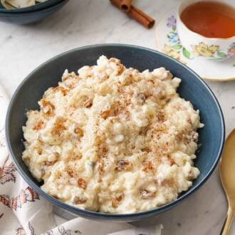 Pinterest graphic of a blue bowl of rice pudding with cinnamon sprinkled on top and a cup of tea in the background.