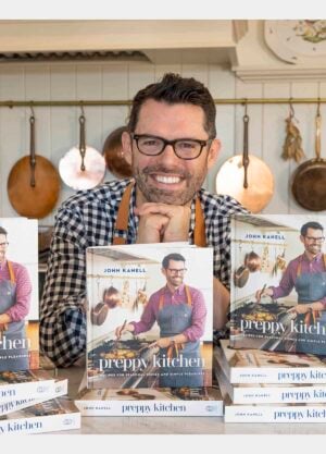 John Kanell in his kitchen behind stacks of the new Preppy Kitchen Cookbook.