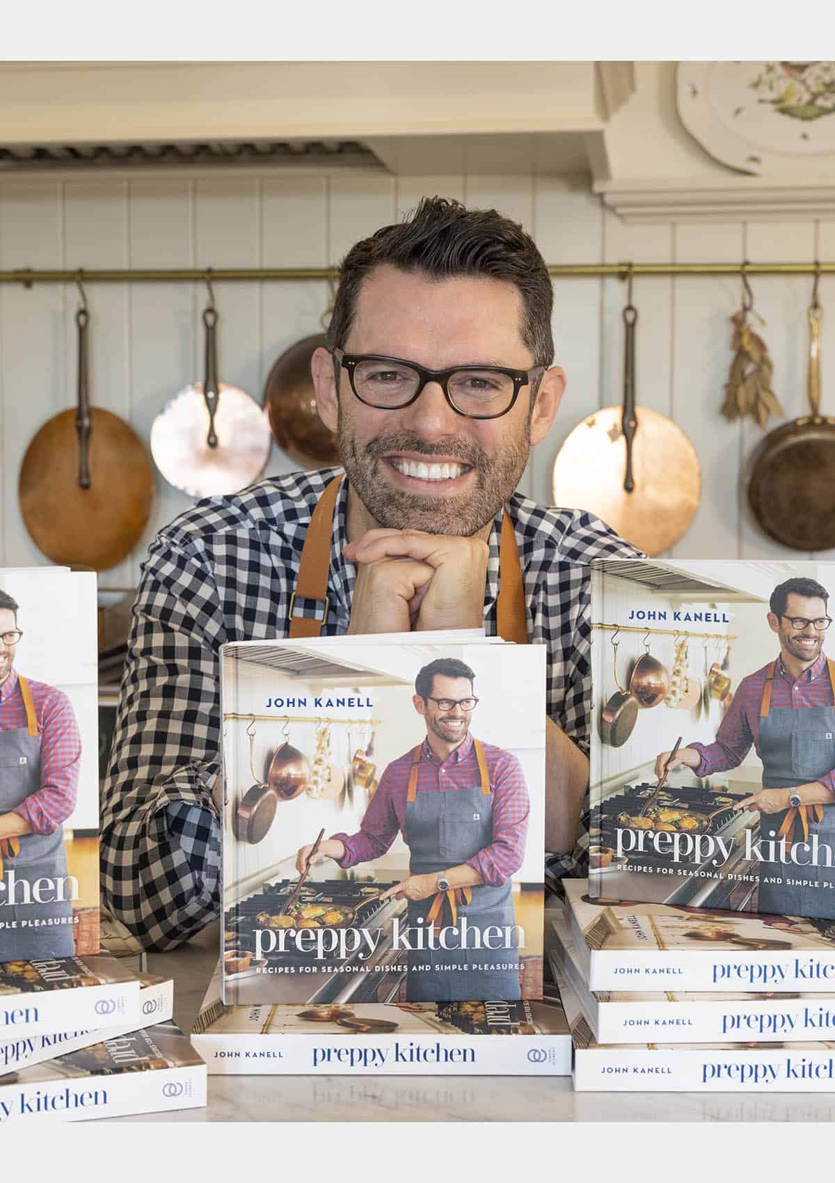 John Kanell in his kitchen behind stacks of the new Preppy Kitchen Cookbook.