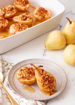 A plate with two baked pears with a baking dish with more in the background and some fresh pears on the side.
