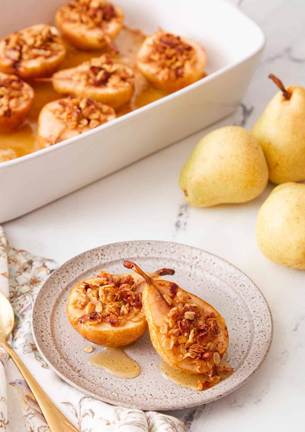 A plate with two baked pears with a baking dish with more in the background and some fresh pears on the side.