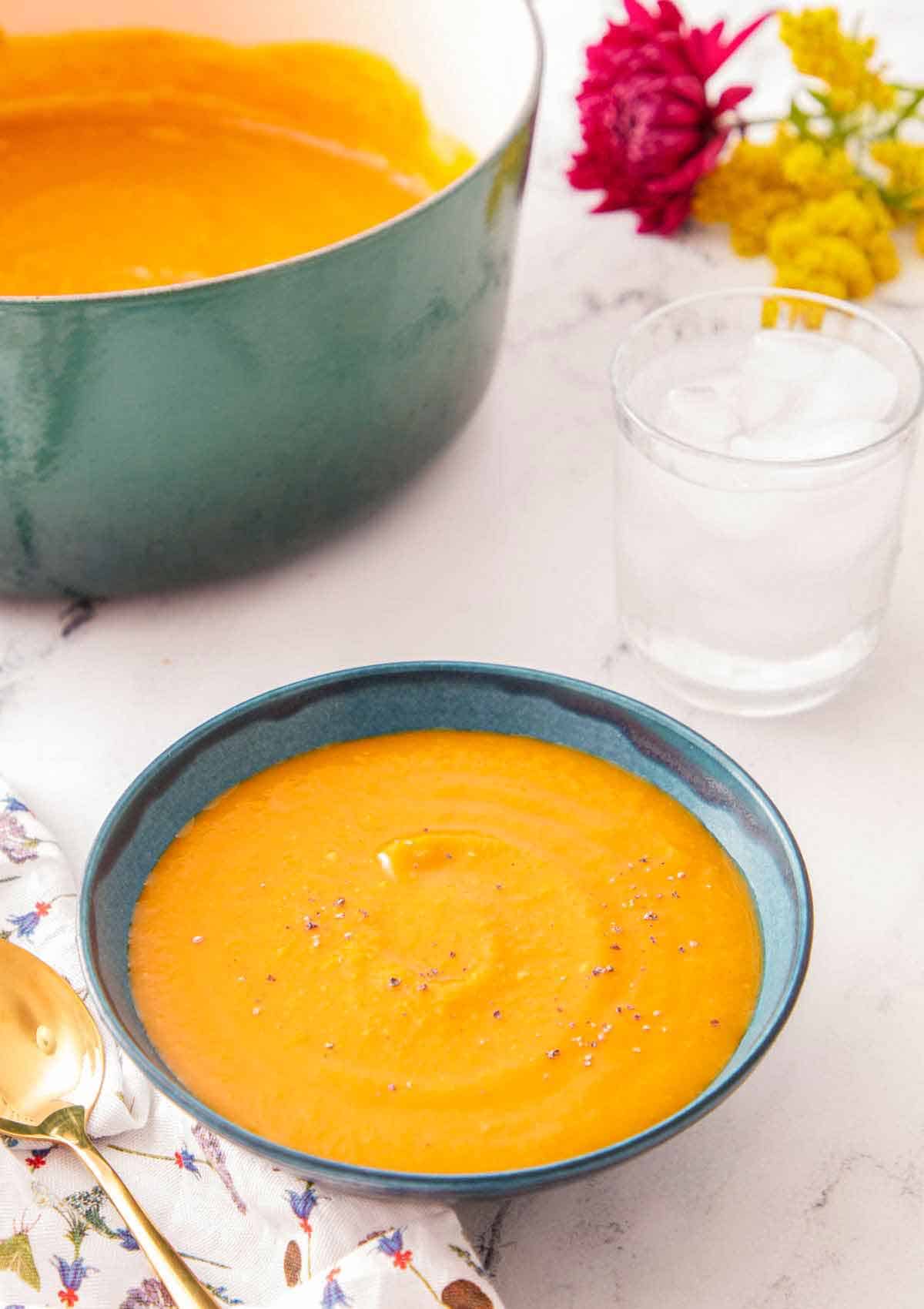 A bowl of butternut squash soup with a glass of water and pot of soup off to the side.