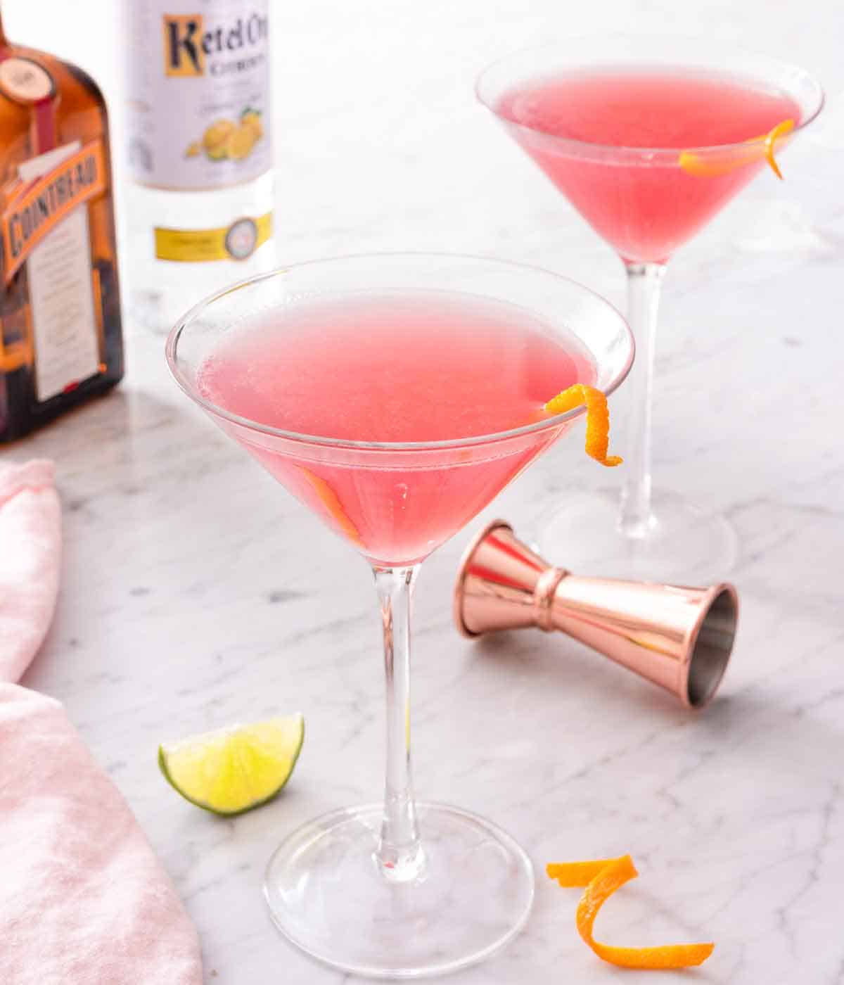 Two glasses of Cosmopolitan with an orange garnish in each glass with bottles of alcohol in the background.
