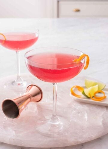 Two glasses of Cosmopolitan with an orange garnish on a marble tray with ice and a jigger with more garnishes.