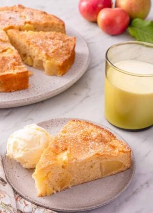 A slice of French apple cake with a scoop of ice cream beside the slice. The rest of the cake, cut, in the background.