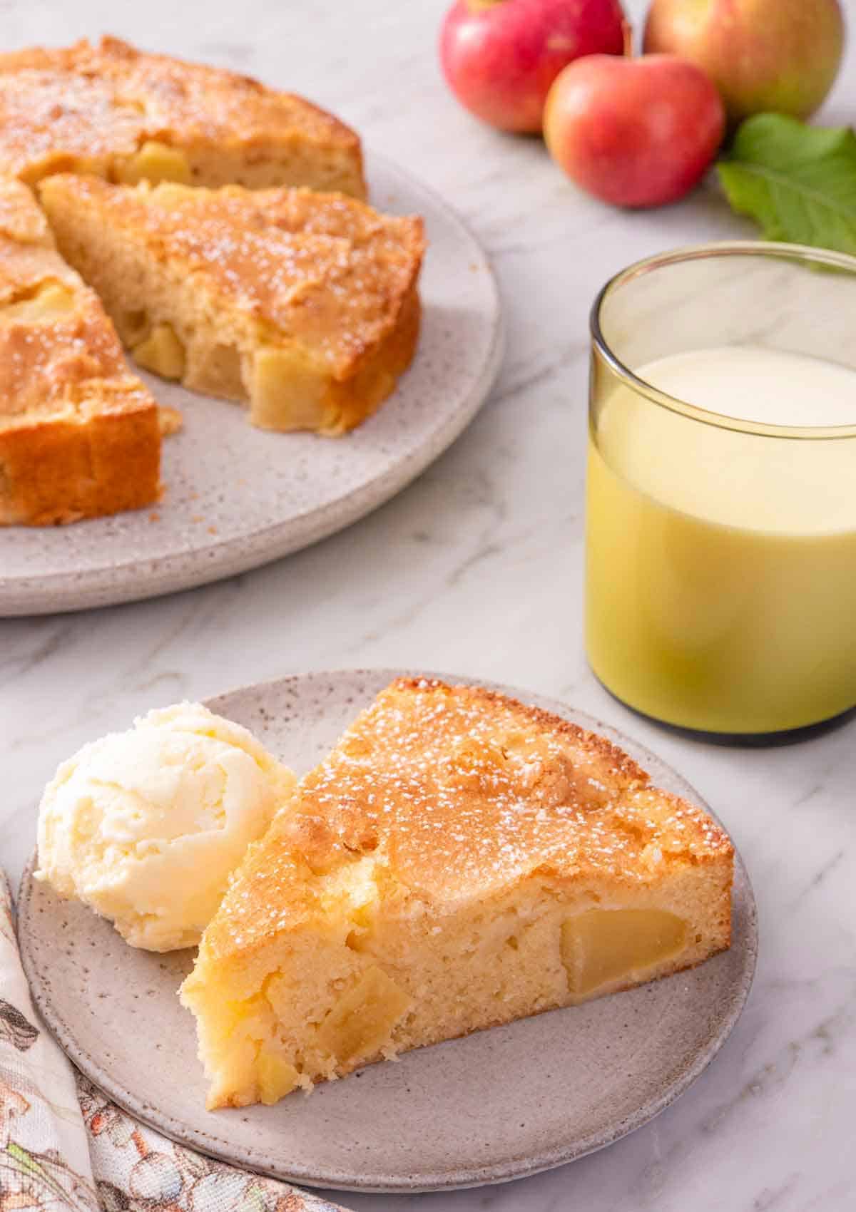 A slice of French apple cake with a scoop of ice cream beside the slice. The rest of the cake, cut, in the background.