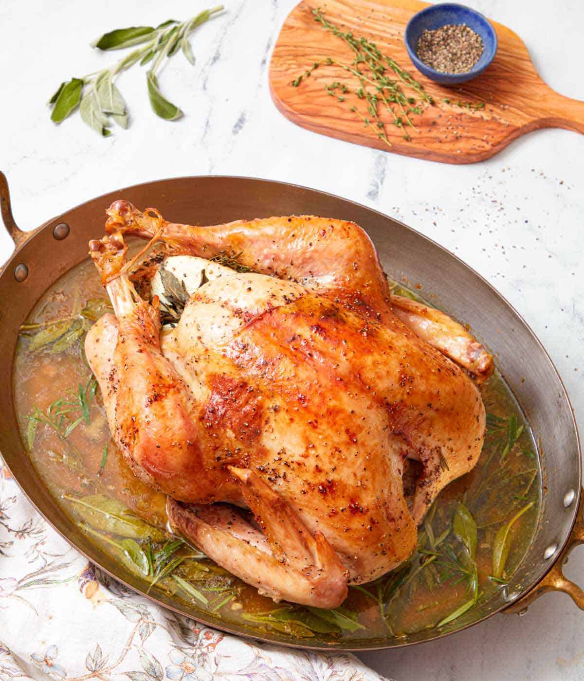 A cooked turkey in a roasting pan overtop the cooking liquid and fresh herbs.