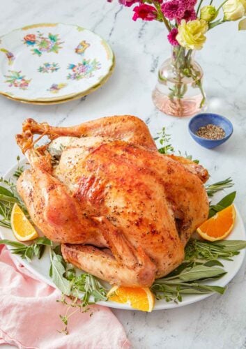 A platter with a roasted turkey over fresh herbs and orange wedges.