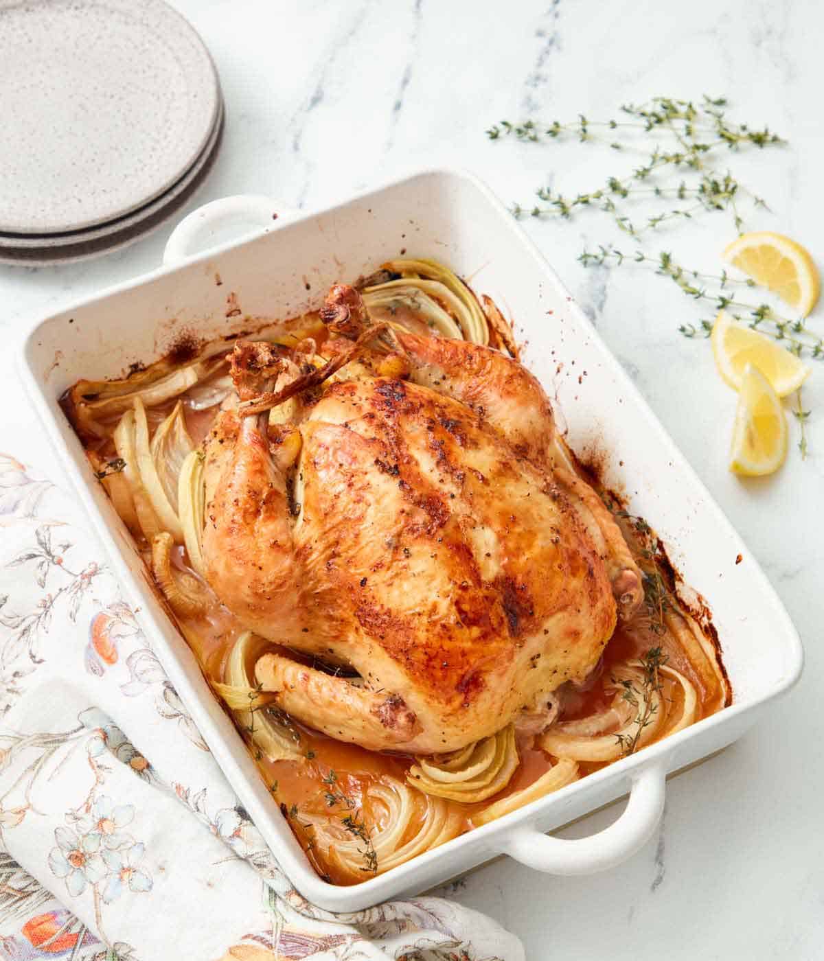 A baking dish with onions and thyme underneath a whole roasted chicken. More thyme and lemons scattered on the side.