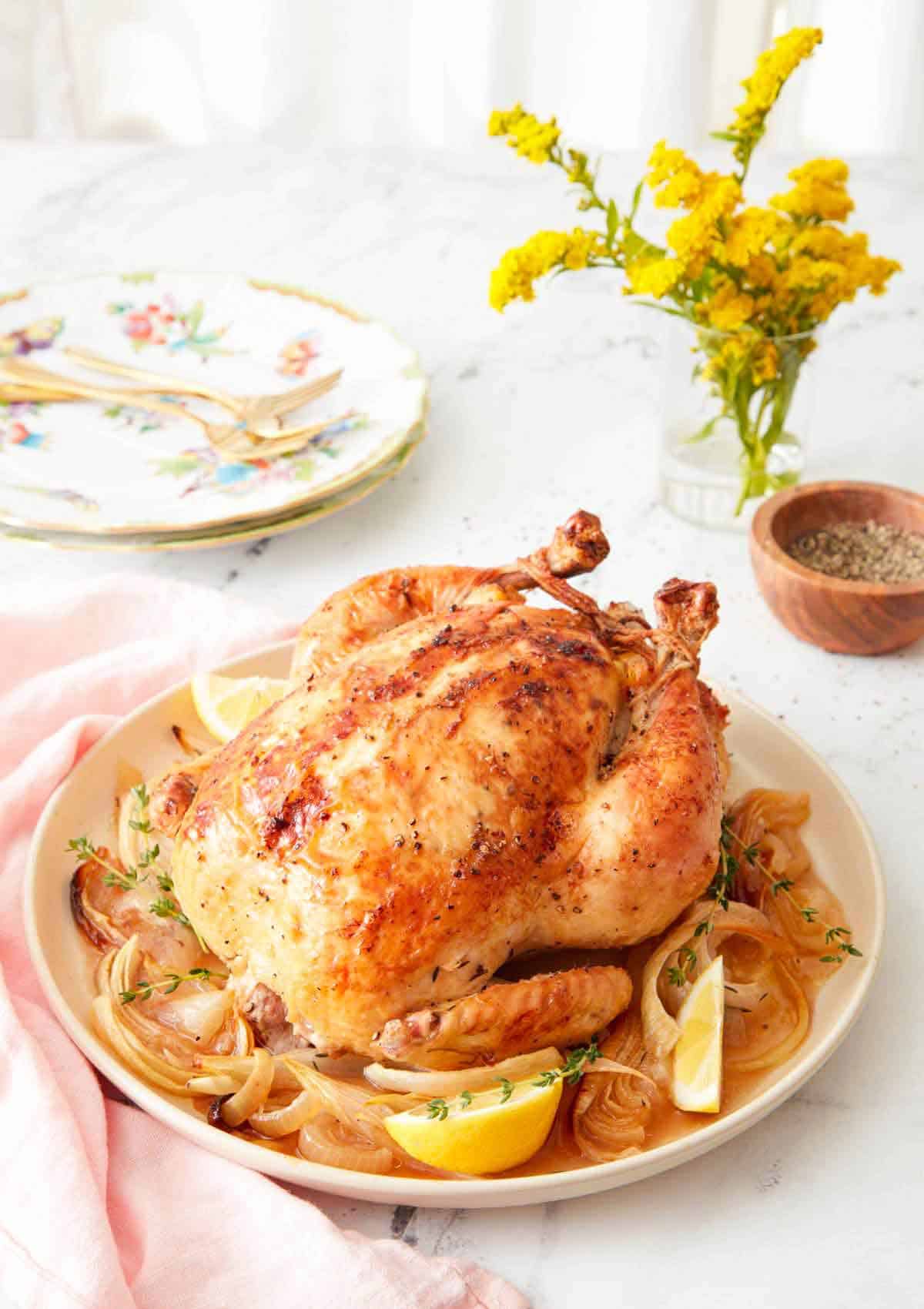 A roasted chicken on a plate of onions, lemon, and thyme. A small bunch of flowers, peppers, and plates in the background.
