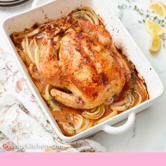 Pinterest graphic of a white baking dish with onions underneath a roasted chicken. Cut lemon and thyme beside it on the counter.