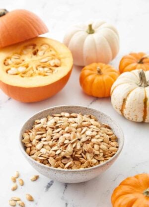 A bowl of roasted pumpkin seeds with a cut pumpkin in the background and small pumpkins scattered around.