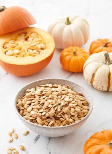 A bowl of roasted pumpkin seeds with a cut pumpkin in the background and small pumpkins scattered around.