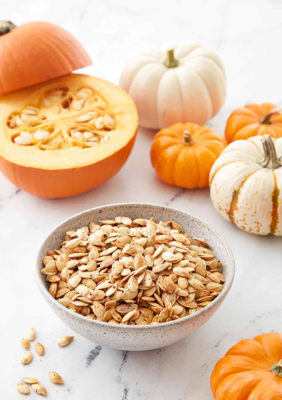 A bowl of roasted pumpkin seeds with a cut pumpkin in the background and small pumpkins scattered around.