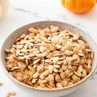 A bowl of roasted pumpkin seeds with a few on the counter and some pumpkins off to the side.