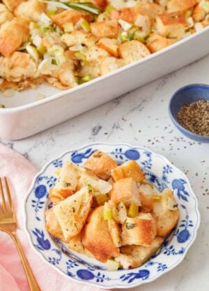A plate of stuffing with a pinch bowl of pepper and the rest of the baking dish of stuffing in the background.