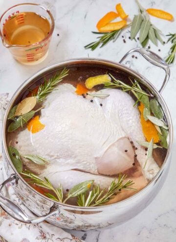 A pot of turkey brine with a large turkey inside with extra ingredients scattered on the counter.
