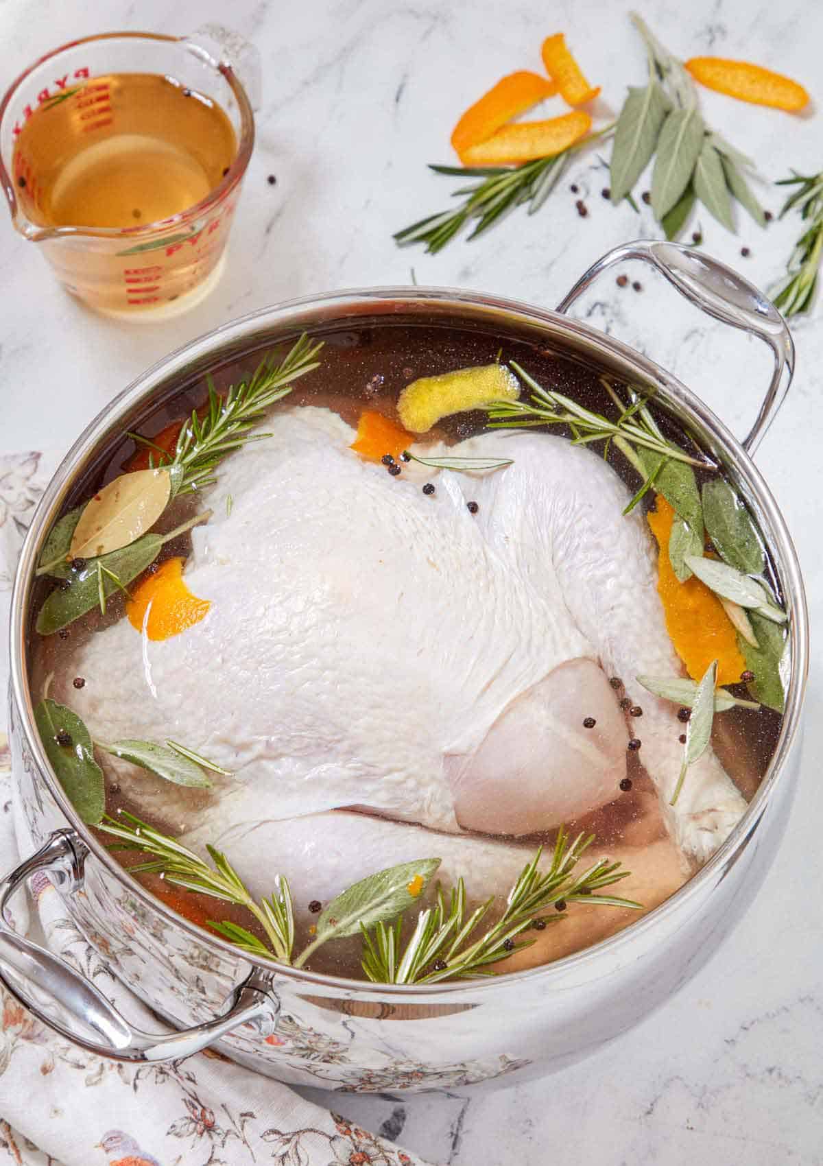 A pot of turkey brine with a large turkey inside with extra ingredients scattered on the counter.