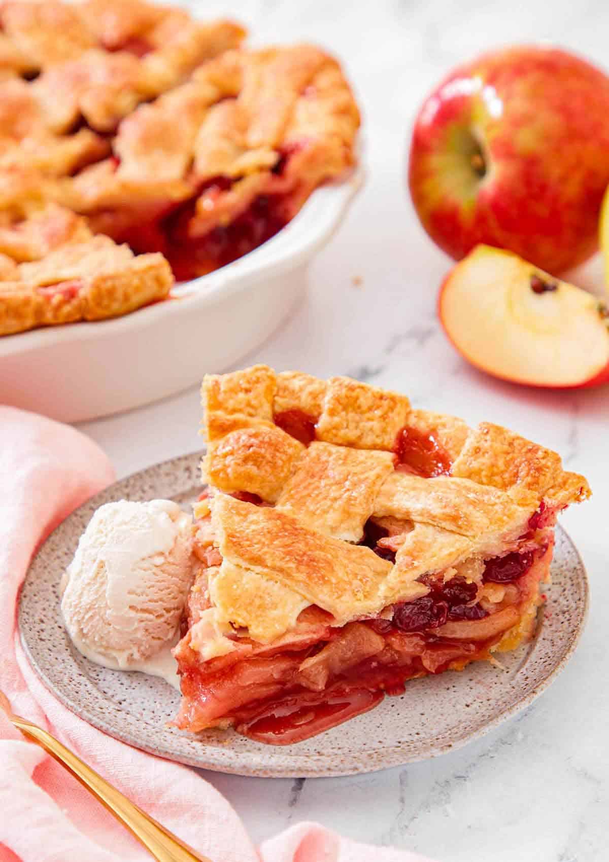 A slice of apple cranberry pie with a scoop of ice cream on the side. Rest of the pie in the background.