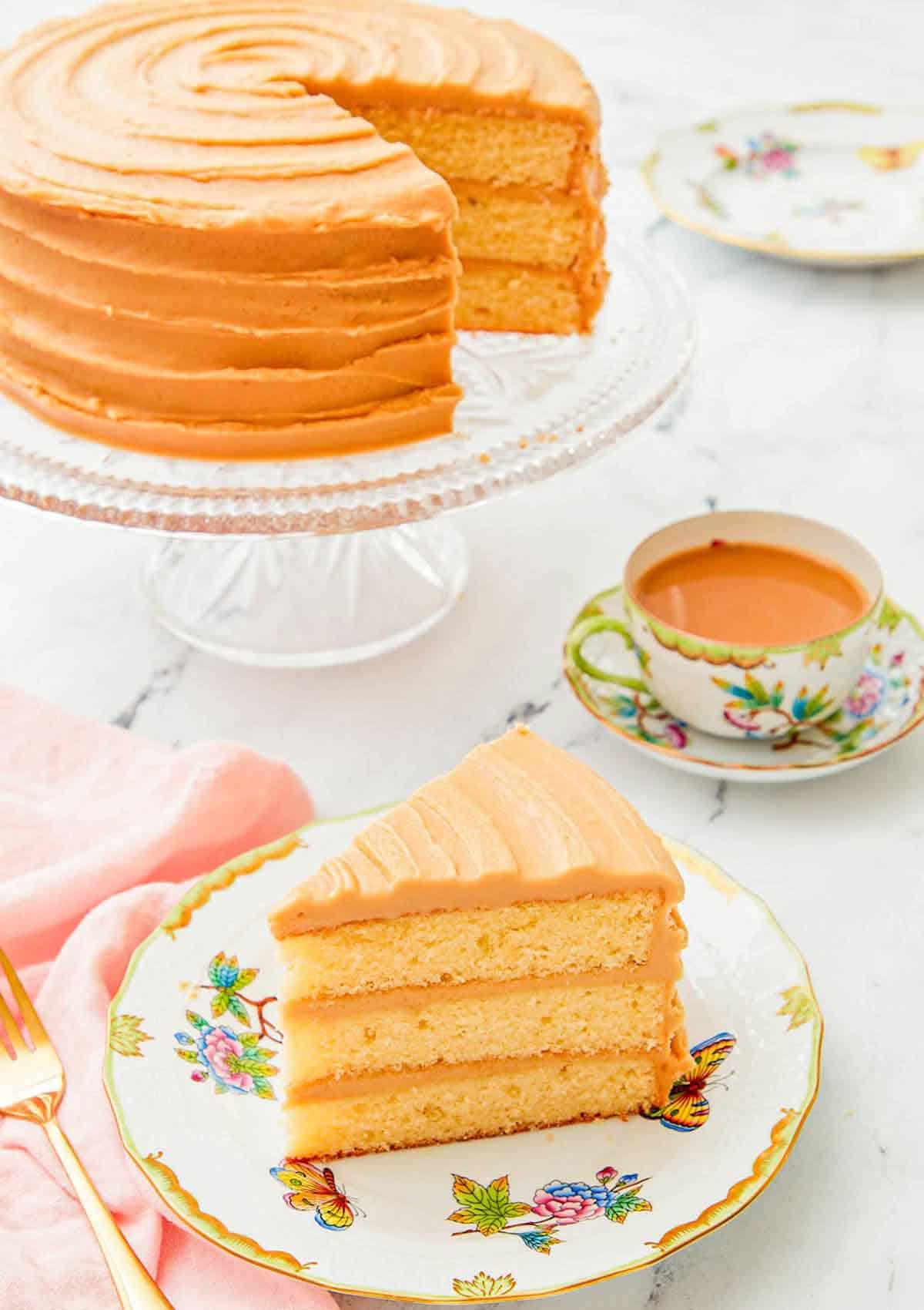 A slice of caramel cake on a plate with the rest of the cake on a stand in the background along with coffee.