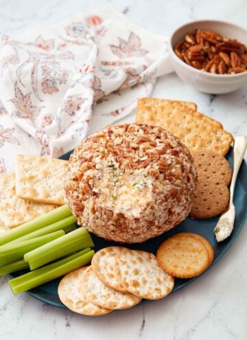 A platter with a cheese ball with a bit scraped off. Crackers and celery on the side. A bowl of pecans in the background.