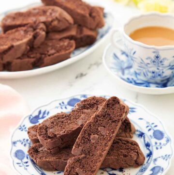 A plate with multiple chocolate biscottis with a cup of coffee and more biscotti in the background.