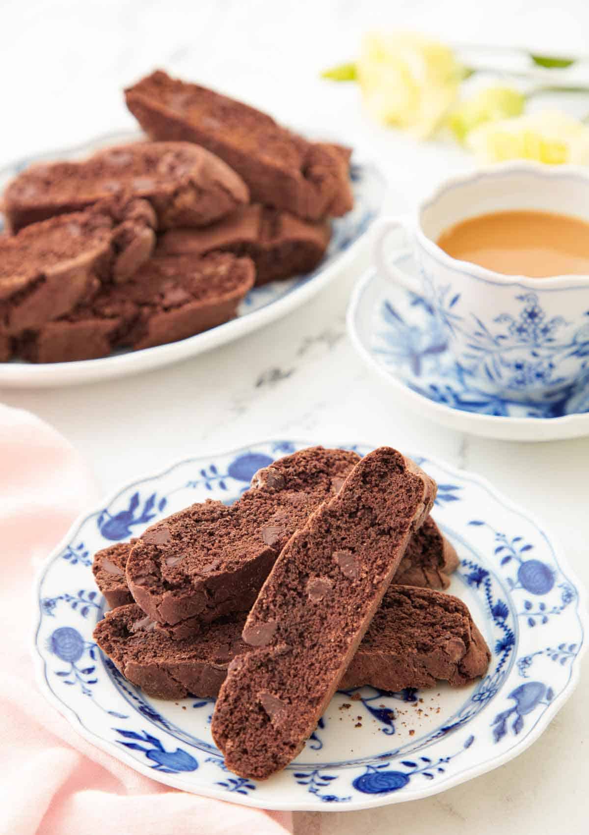 A plate with multiple chocolate biscottis with a cup of coffee and more biscotti in the background.