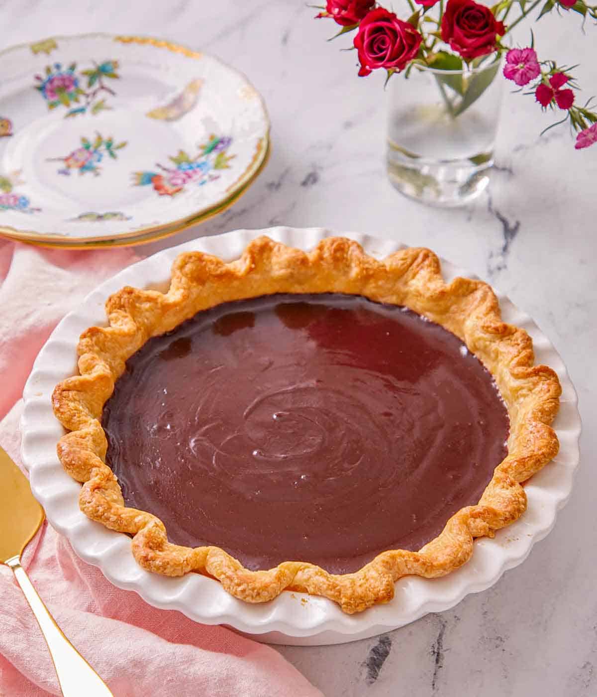A chocolate pie in a white baking dish with flowers and a stack of plates in the background.