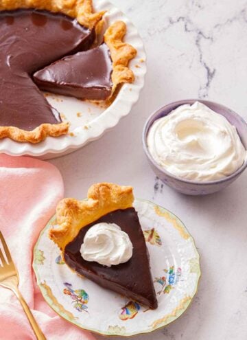 A slice of chocolate pie with a bowl of whipped cream and the rest of the pie in the background.