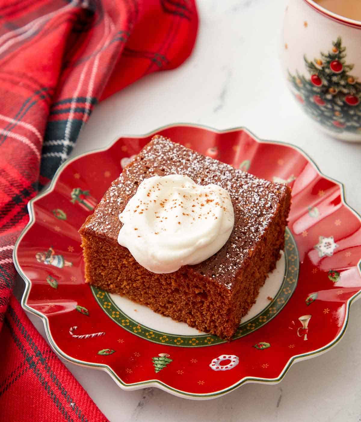 Overhead view of a festive red plate with a square slice of gingerbread cake with a dollop of whipped cream on top.