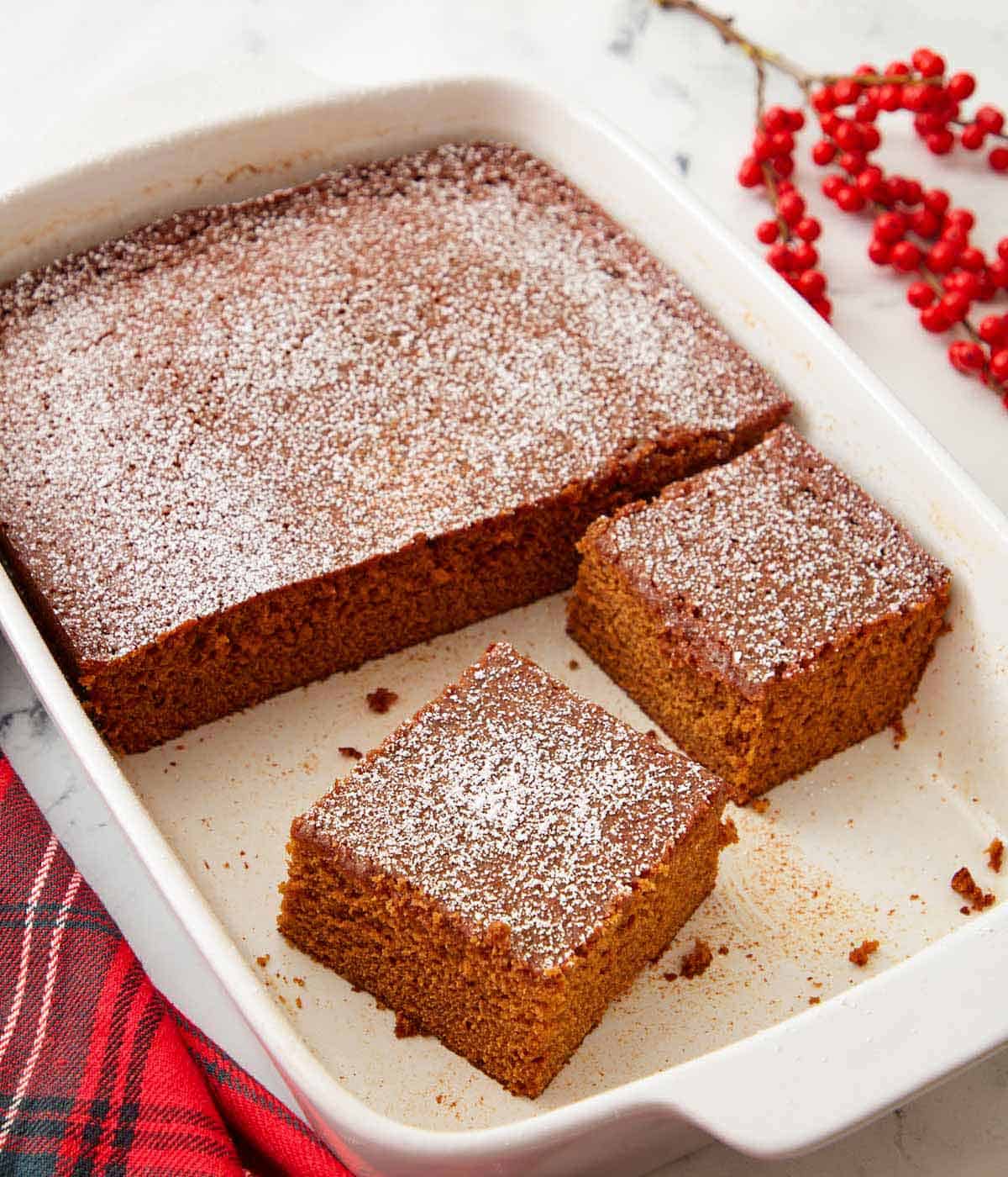 A white baking dish containing half of a gingerbread cake with two square slices cut.