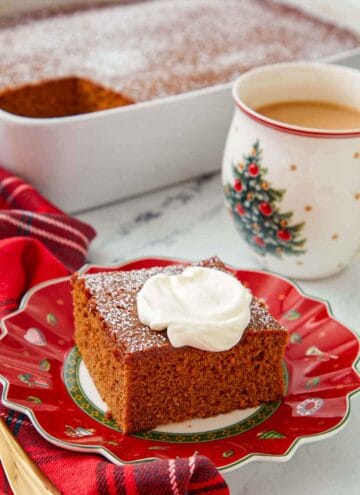 A festive red plate with a square slice of gingerbread cake with a dollop of whipped cream on top with a cup of coffee and the rest of the cake in the background.
