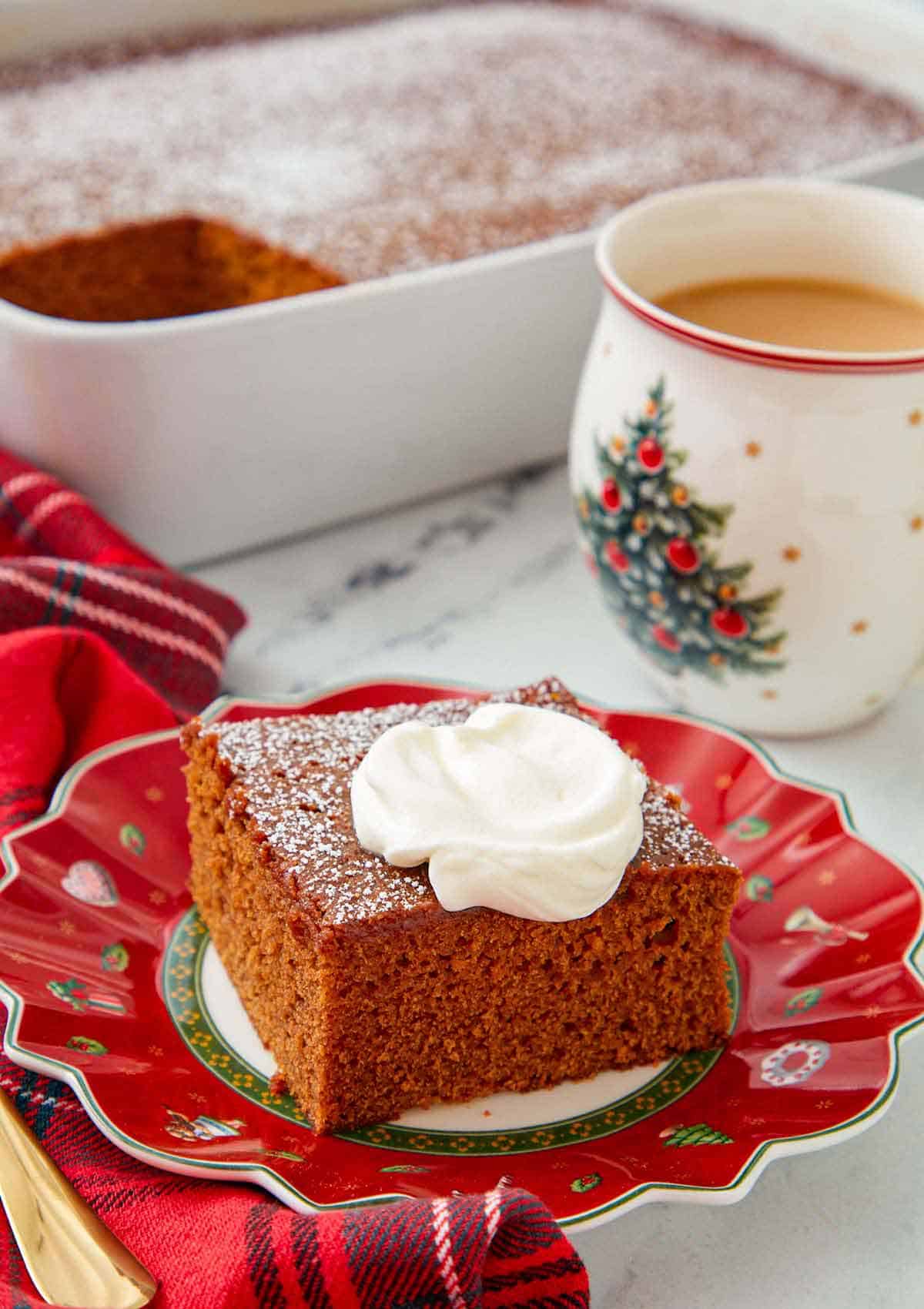 A festive red plate with a square slice of gingerbread cake with a dollop of whipped cream on top with a cup of coffee and the rest of the cake in the background.