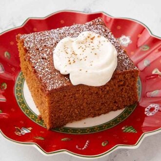 A red festive plate with a slice of gingerbread cake with a dusting of powdered sugar and dollop of whipped cream on top.