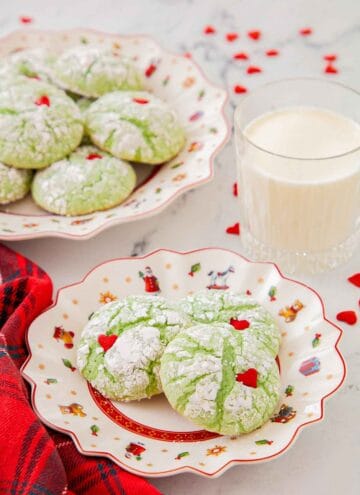 A plate with three Grinch cookies with a glass of milk and additional cookies in the background.