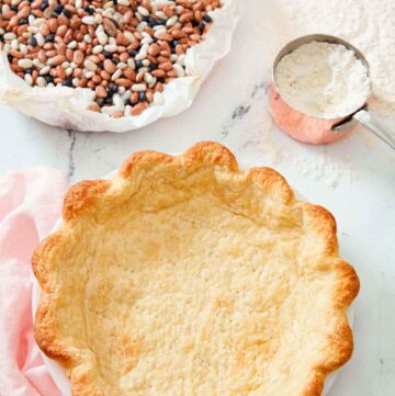 A baked pie crust with dried beans in parchment and a measuring cup of flour in the background.