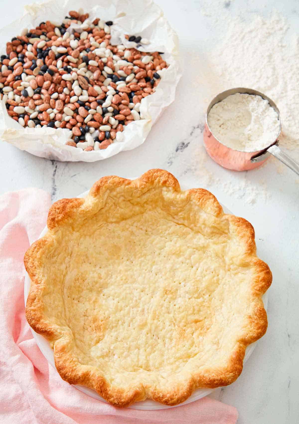 A baked pie crust with dried beans in parchment and a measuring cup of flour in the background.