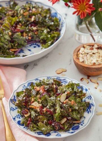A plate of kale salad with a small bowl of almonds and serving bowl of salad in the back.
