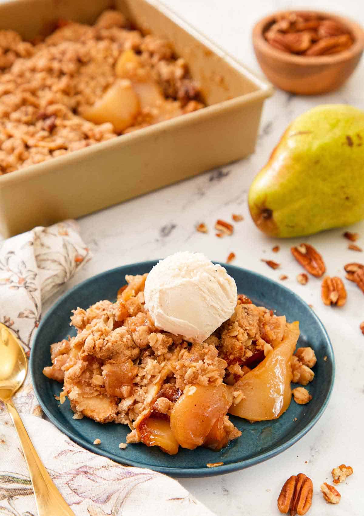 A plate of pear crisp with a scoop of vanilla ice cream on top with the rest of the baking dish in the back alongside a pear.
