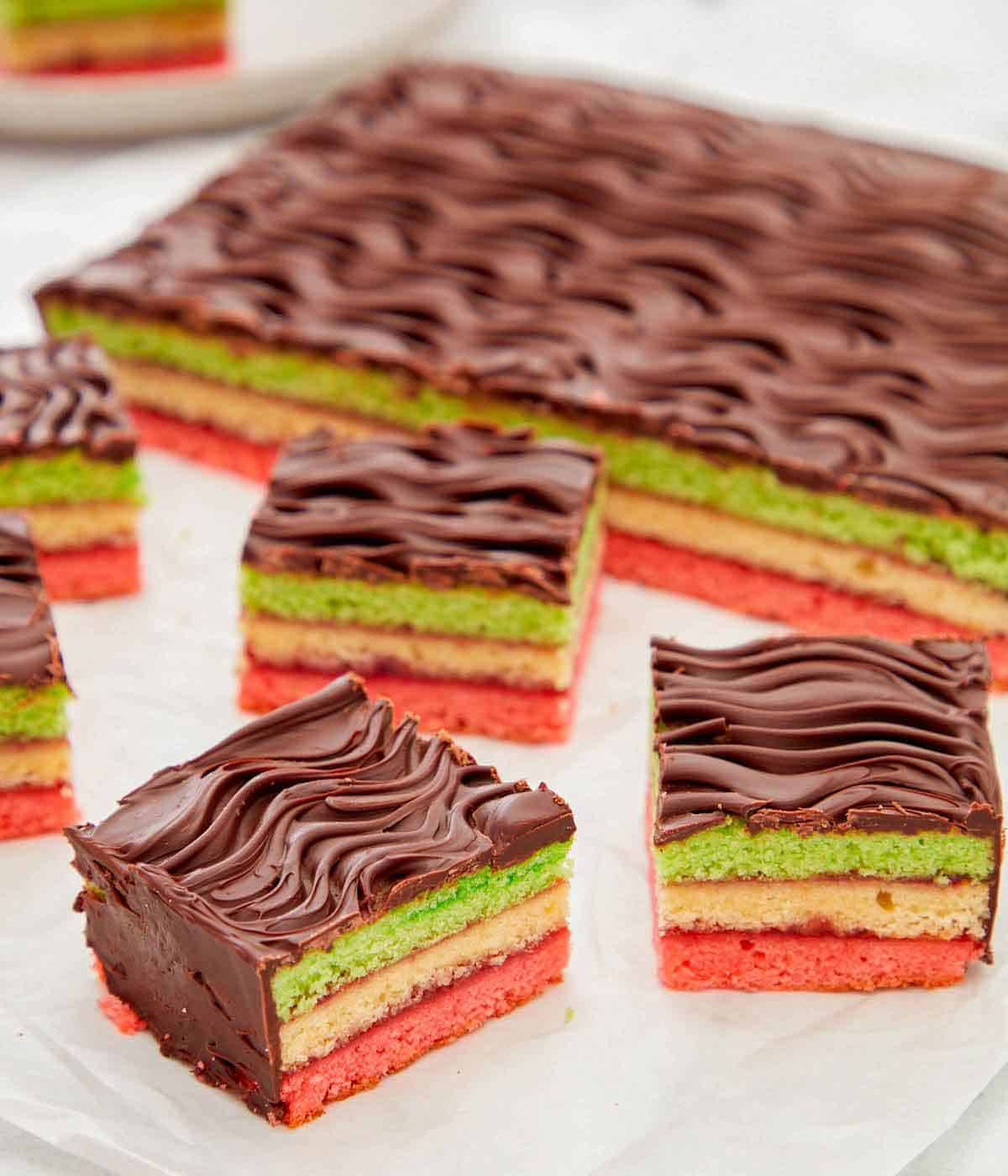 Multiple pieces of rainbow cookies cut from the main dessert in the background.
