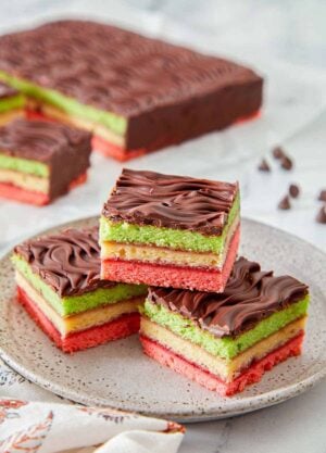 A plate with three pieces of rainbow cookies in a pyramid. The rest of the dessert in the background.