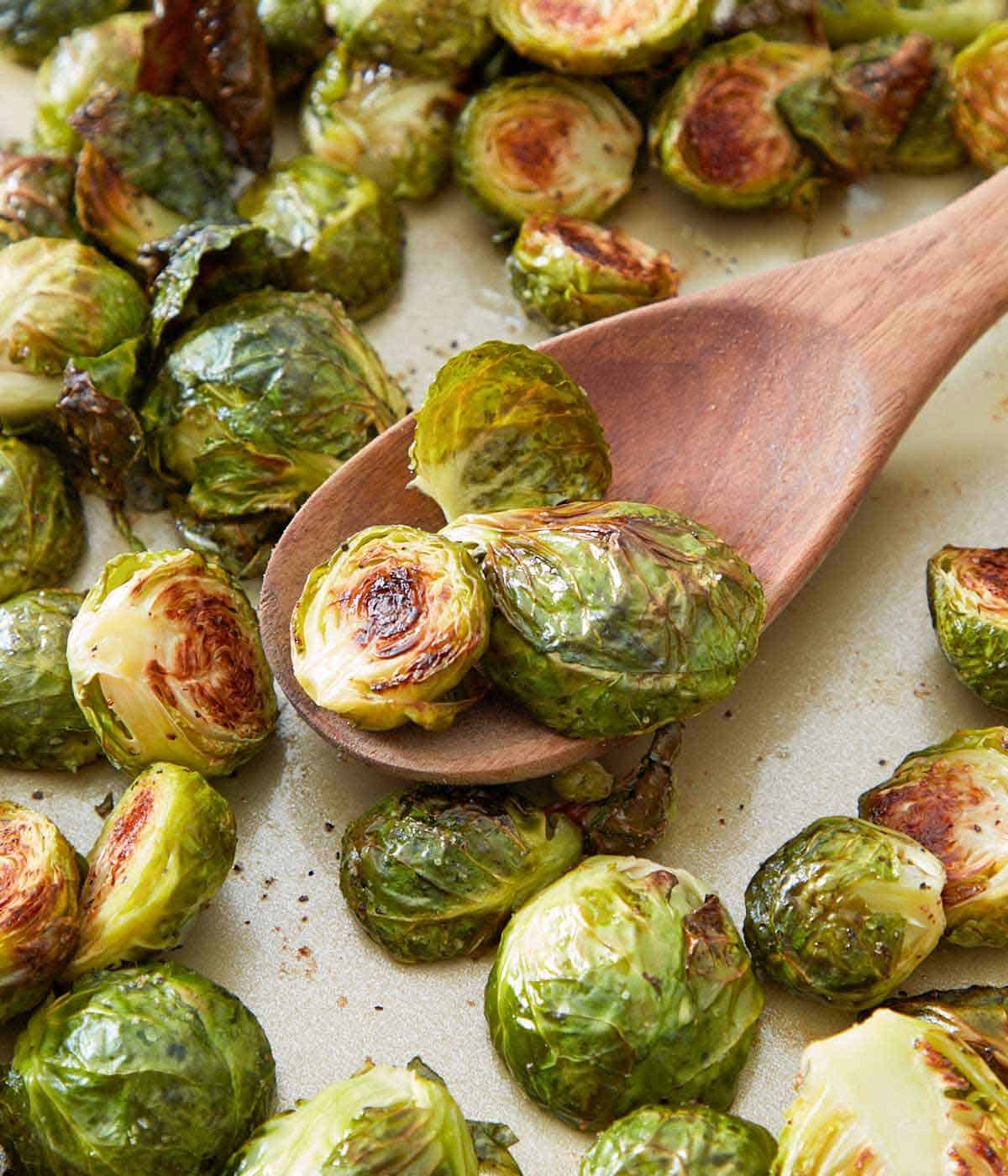 A wooden spoon with some roasted Brussels sprouts surrounded by more sprouts.