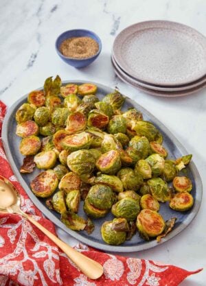 A blue oval serving platter with roasted Brussels sprouts with a spoon, stack of plates, and pepper on the side.