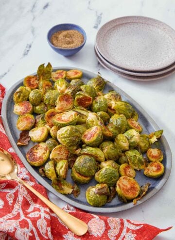 A blue oval serving platter with roasted Brussels sprouts with a spoon, stack of plates, and pepper on the side.