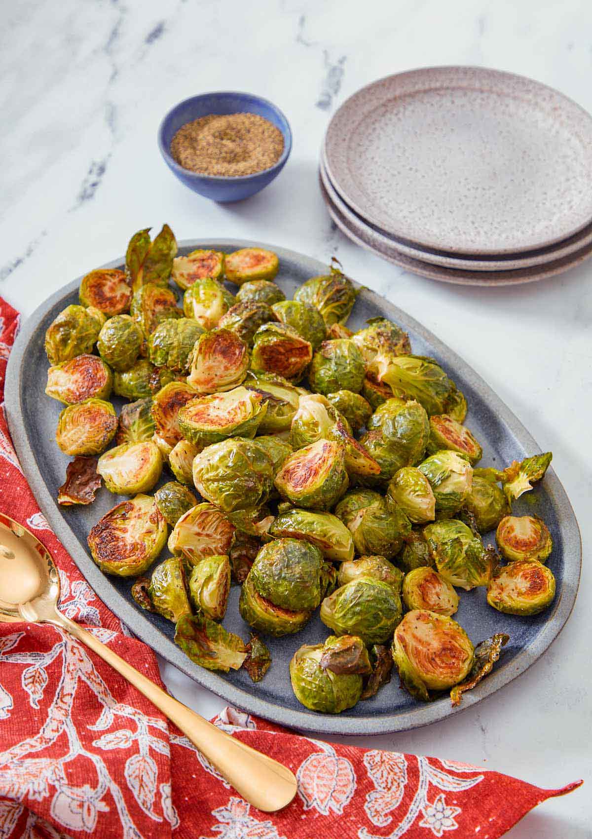 A blue oval serving platter with roasted Brussels sprouts with a spoon, stack of plates, and pepper on the side.