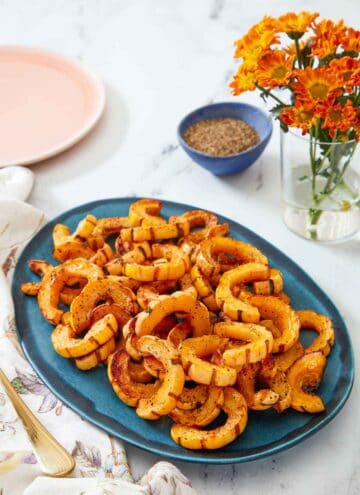 A blue serving platter with roasted delicata squash. A vase of flowers and a pinch bowl of pepper in the background.